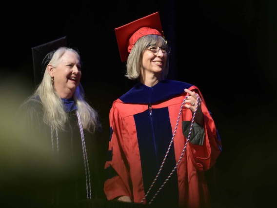 Two University of Arizona College of Nursing faculty dressed in graduation caps and gowns stand on a stage smiling as they look off to the side. 