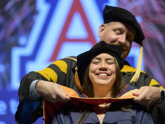 A University of Arizona College of Nursing professor places a hood over the shoulders of a nursing student. Both are dressed in graduation caps and gowns.