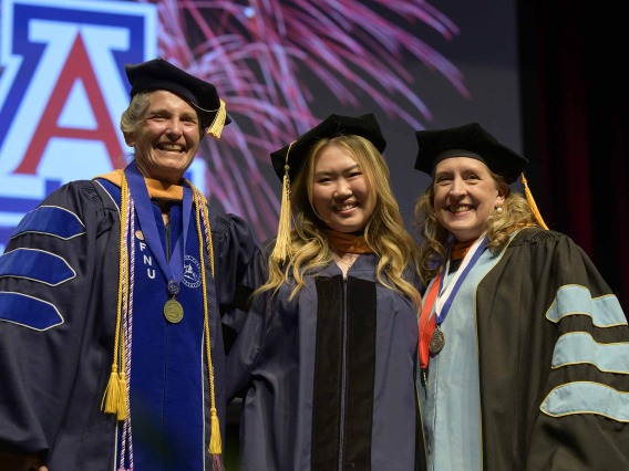 Two University of Arizona College of Nursing professor flank a student on stage. All are wearing graduation caps and gowns. 