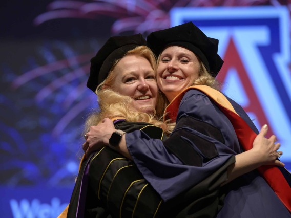 A University of Arizona College of Nursing professor hugs a nursing student. Both are dressed in graduation caps and gowns.