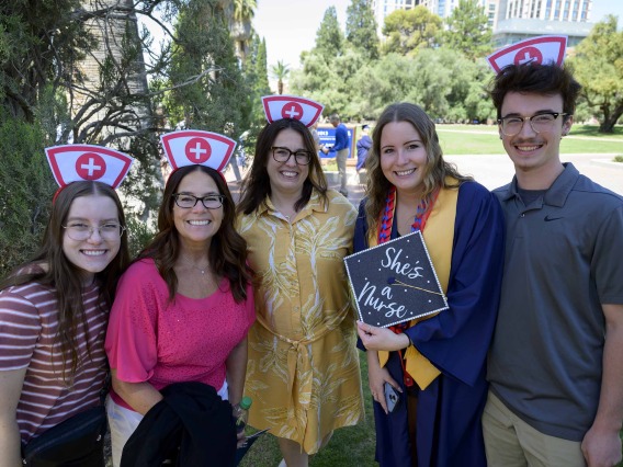 A family stands on either side of a recent graduate of the University of Arizona College of Nursing who is dressed in a graduation cap and gown. The family members have white paper hats with a red cross on them. 