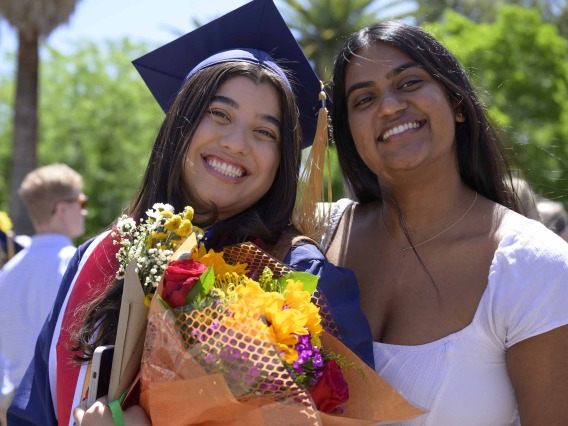 A University of Arizona College of Nursing student in a cap and gown holding flowers stands next to her friend outside. Both are smiling. 