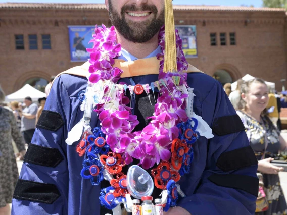 A University of Arizona College of Nursing doctor of nursing practice graduate stands outside smiling in a graduation cap and gown. He has flowers around his neck and a necklace made out of a variety of medical supplies used in his profession. 