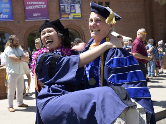 A University of Arizona College of Nursing professor carries a Nurse-Midwifery graduate. Both are dressed in graduation caps and gowns and laughing. 