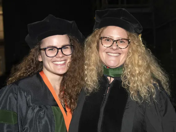 Two women, a mother and daughter, both in graduation caps and gowns, stand together, smiling, before the University of Arizona College of Medicine – Tucson commencement ceremony. 