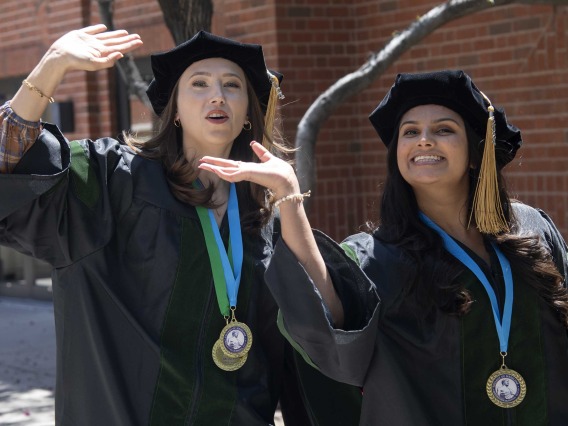 Two University of Arizona College of Medicine – Tucson students in graduation regalia wave as they walk outside on their way to their graduation ceremony.