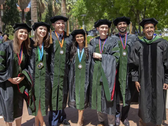 Six University of Arizona College of Medicine – Tucson students and one professor, all in graduation regalia, stand together outside before going to their commencement ceremony. 