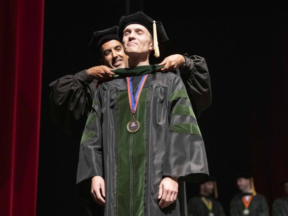 A University of Arizona College of Medicine – Tucson student dressed in a graduation cap and gown has a ceremonial hood placed over his shoulders by his faculty mentor, who is also dressed in graduation regalia.