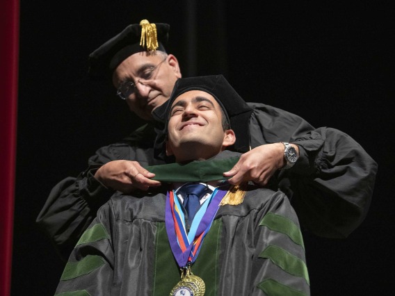A University of Arizona College of Medicine – Tucson student dressed in a graduation cap and gown has a ceremonial hood placed over his shoulders by his father, who is also dressed in graduation regalia.