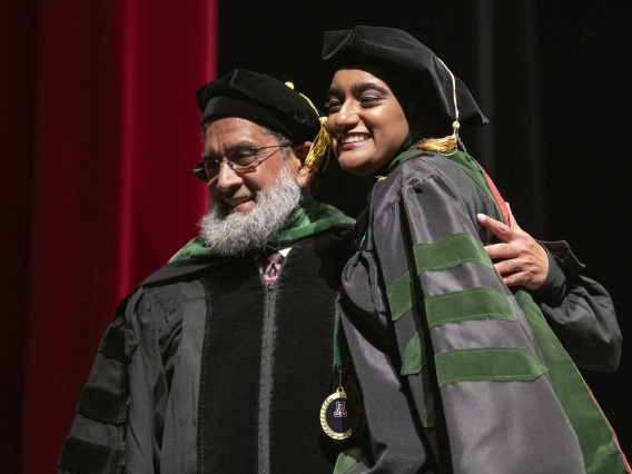 A University of Arizona College of Medicine – Tucson student dressed in a graduation cap and gown hugs her father, who is also dressed in graduation regalia, while standing on a stage.