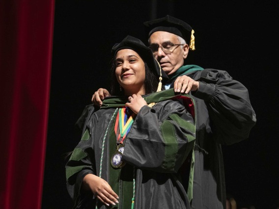 A University of Arizona College of Medicine – Tucson student dressed in a graduation cap and gown has a ceremonial hood placed over her shoulders by her father, who is also dressed in graduation regalia. 
