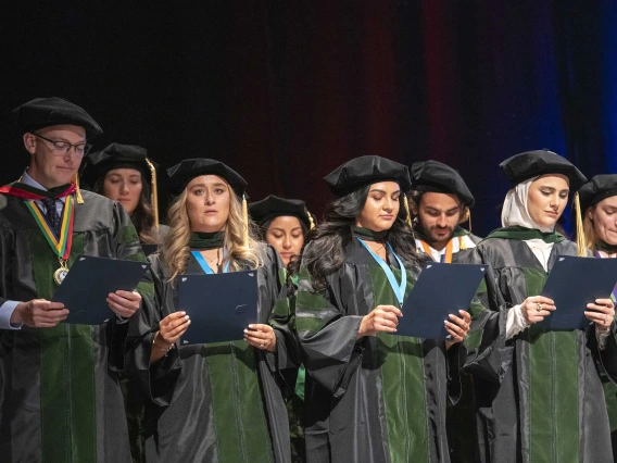 A large group of University of Arizona College of Medicine – Tucson graduates dressed in caps and gowns stand on a stage reading the Hippocratic Oath. 