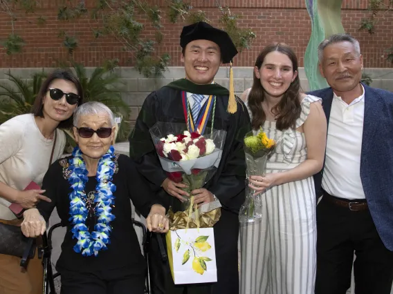 A University of Arizona College of Medicine – Tucson graduate smiles as he holds flowers and a gift bag while surrounded by family and friends. 