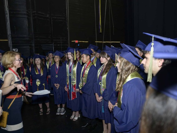 A large group of University of Arizona R. Ken Coit College of Pharmacy students wearing graduation caps and gowns stands together listening to a staff member.