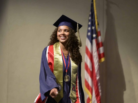 A University of Arizona R. Ken Coit College of Pharmacy student dressed in a graduation cap and gown smiles as she walks in front of an American flag. 