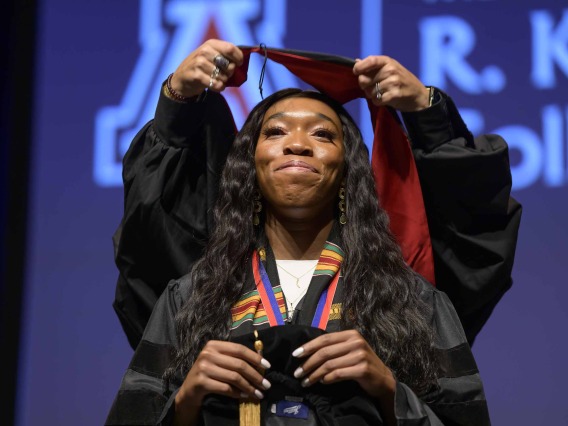A University of Arizona R. Ken Coit College of Pharmacy student in a graduation gown smiles as a professor places a graduation hood over her head.