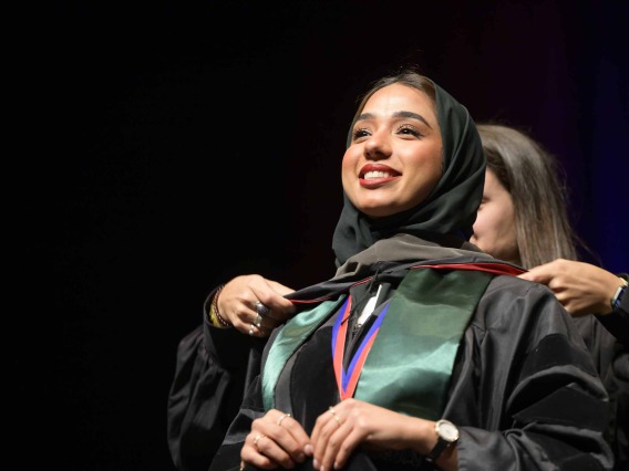 A University of Arizona R. Ken Coit College of Pharmacy Doctor of Pharmacy student wearing a graduation gown has a ceremonial hood placed over her shoulders as she smiles. 