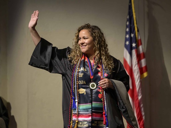A University of Arizona R. Ken Coit College of Pharmacy student dressed in a graduation gown waves and smiles. There is an American flag behind her. 