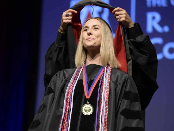 A University of Arizona R. Ken Coit College of Pharmacy student in a graduation gown smiles as a professor places a graduation hood over her head.