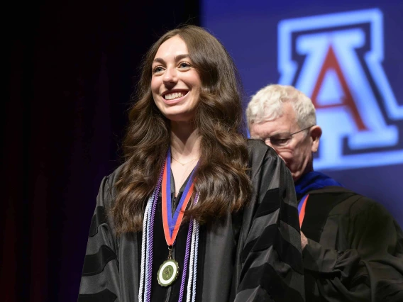 A University of Arizona R. Ken Coit College of Pharmacy student in a graduation gown smiles as a professor prepares to place a graduation hood over her head.