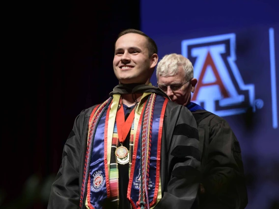 A University of Arizona R. Ken Coit College of Pharmacy student in a graduation gown smiles as a professor prepares to place a graduation hood over his head.