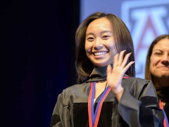 A University of Arizona R. Ken Coit College of Pharmacy student dressed in a graduation gown waves and smiles as a faculty member smiles behind her.