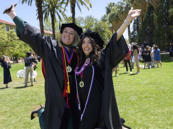 Two University of Arizona R. Ken Coit College of Pharmacy students wearing graduation caps and gowns celebrate with their hands in the air.