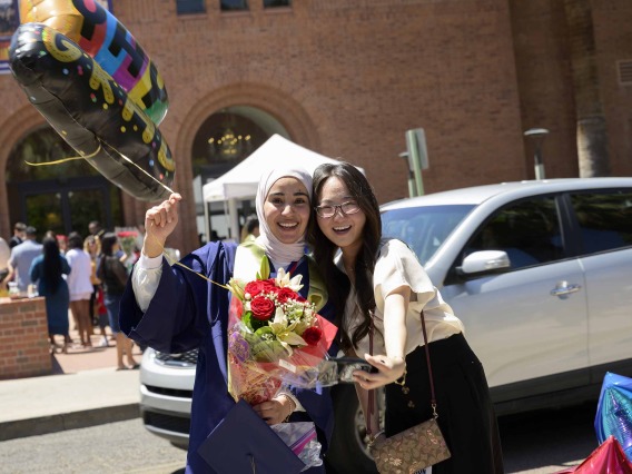 Two University of Arizona R. Ken Coit College of Pharmacy students, one wearing a graduation gown and holding a balloon and flowers, hug after the college’s graduation ceremony.