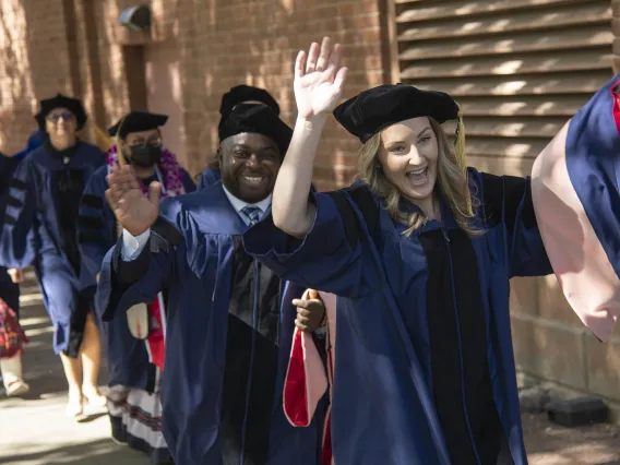 Several University of Arizona Mel and Enid Zuckerman College of Public Health students wearing graduation caps and gowns smile and wave as they walk outside. 