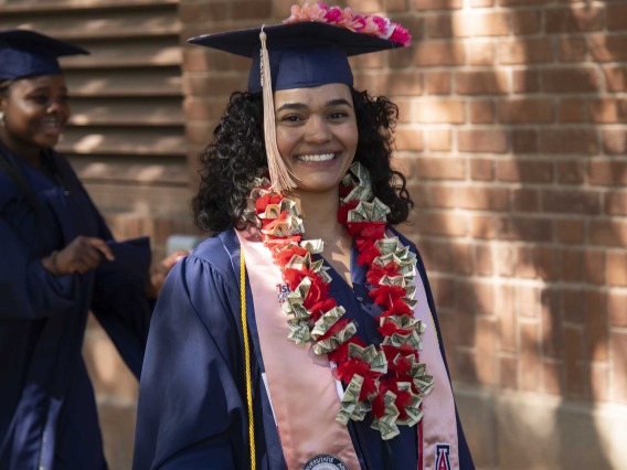 A University of Arizona Mel and Enid Zuckerman College of Public Health student wearing a graduation cap and gown smiles while walking outside. 