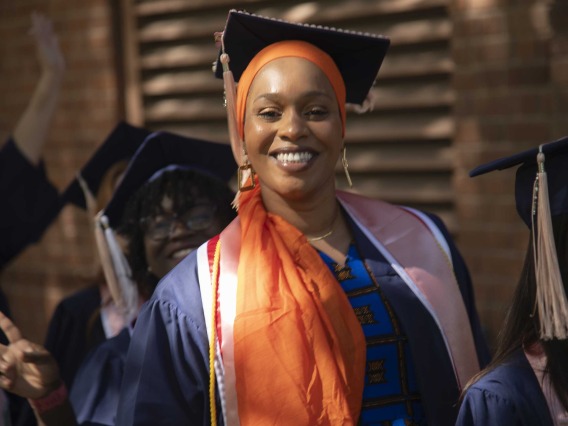 A smiling University of Arizona Mel and Enid Zuckerman College of Public Health student wearing a graduation cap and gown and an orange headscarf walks outside with other students.