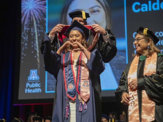 A University of Arizona Mel and Enid Zuckerman College of Public Health student makes a heart sign with her hands as she is hooded by a faculty member as another watches. All are wearing graduation regalia.