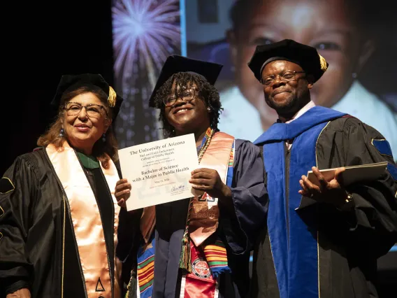 A University of Arizona Mel and Enid Zuckerman College of Public Health student holds up her diploma while standing between two faculty members. All are wearing graduation regalia. 