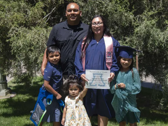 A University of Arizona Mel and Enid Zuckerman College of Public Health student wearing a graduation gown holds up her diploma while standing with her family outside. 