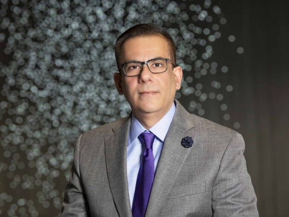 Portrait of a man wearing a grey suit, purple tie and glasses standing in front of lights in the Health Sciences Innovation Building Forum.
