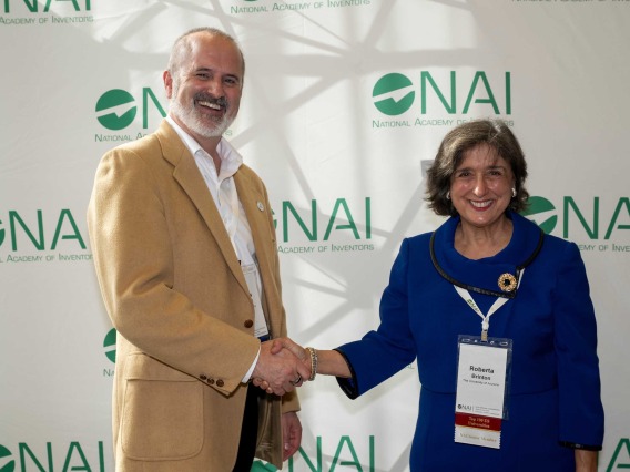 Doug Hockstad, wearing a tan sport coat shakes hands with Dr. Roberta Diaz Brinton as both smile in front of a backdrop that says NAI. 