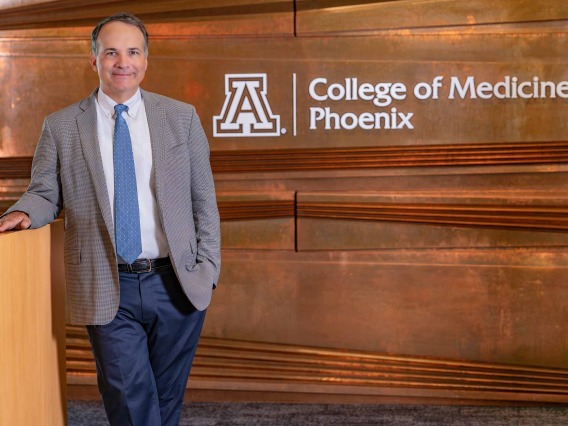 A man in a suit coat stands in front of a wall that has College of Medicine – Phoenix lettering