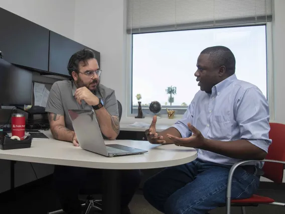 Two men sit in an office talking with an open laptop in front of them.