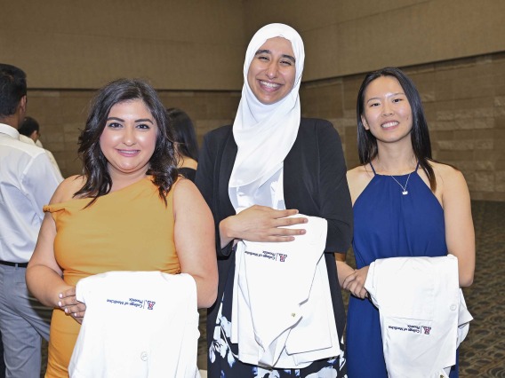 Three new University of Arizona College of Medicine – Phoenix students, with medical white coats draped over their arms, smile as they stand side by side.