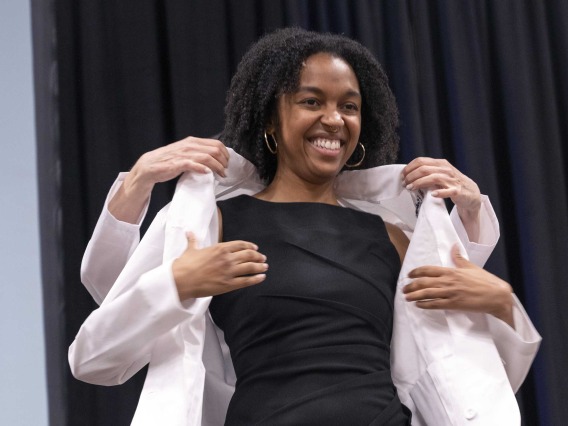 A smiling University of Arizona College of Medicine – Phoenix student in a black dress adjusts a medical white coat that is being put on her by a professor.
