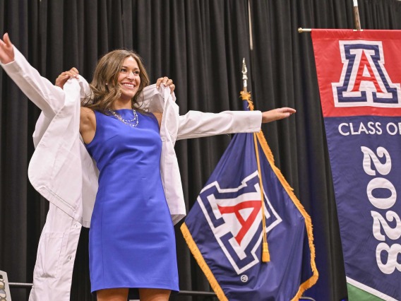 A new University of Arizona College of Medicine – Phoenix student in a blue dress stretches out her arms as a professor puts a medical white coat on her. 