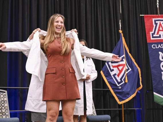 A new University of Arizona College of Medicine – Phoenix student in a brown dress stretches out her arms as a professor puts a medical white coat on her.