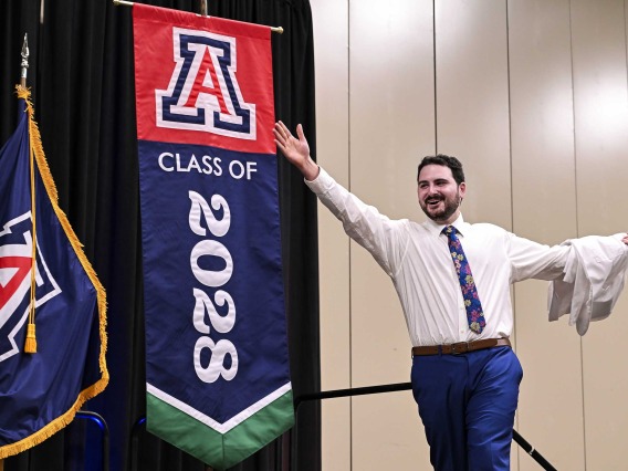 A new University of Arizona College of Medicine – Phoenix student with a medical white coat draped over one arm extends the other arm outward as he smiles and walks onto a stage. Behind him is a  U of A banner that reads “Class of 2028.” 