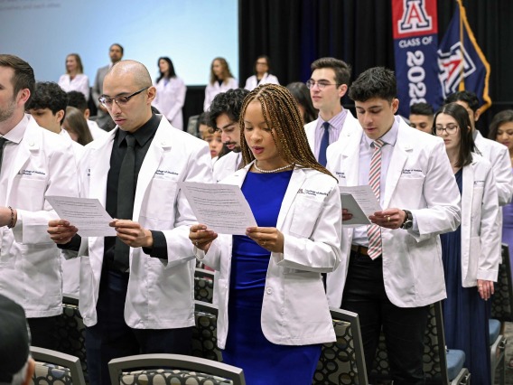 A large group of University of Arizona College of Medicine – Phoenix students stand in their white coats reading from printouts they are holding. 