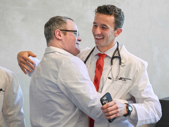 Two University of Arizona College of Medicine – Phoenix students wearing medical white coats smile as they hug. 