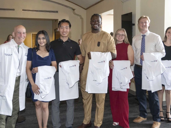 A University of Arizona College of Medicine – Tucson professor wearing a white medical coat stands with six new medical students, all with white coats draped over their arms. 