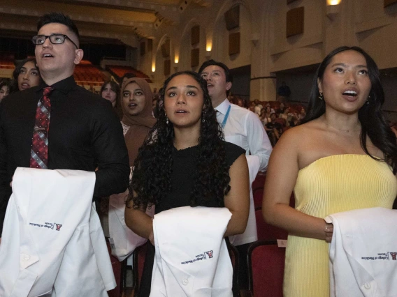 Three new University of Arizona College of Medicine – Tucson students with white coats draped over their arms stand in front of a large group of fellow students and recite their class oath.