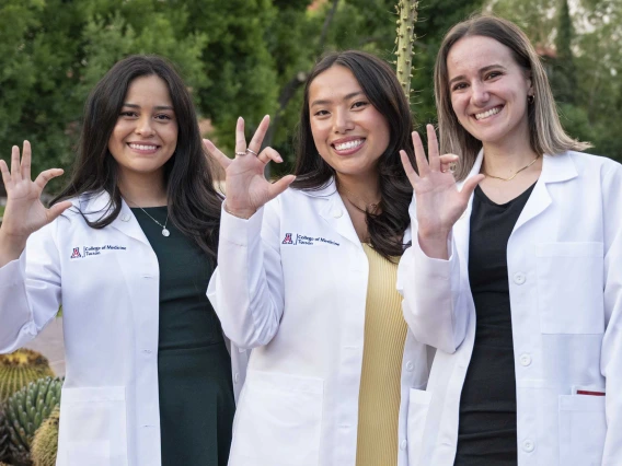 Three new University of Arizona College of Medicine – Tucson students wearing medical white coats smile as they flash the Arizona Wildcats hand sign.