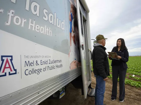 A University of Arizona Mel and Enid Zuckerman College of Public Health professor interviews a farmworker as they stand in a field next to a mobile health trailer. 