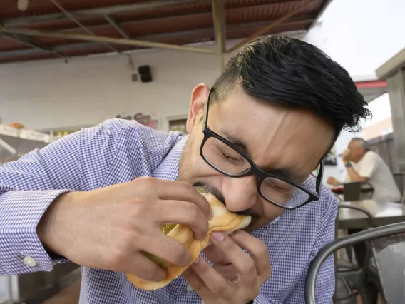 A medical student in a blue shirt bites into a Sonoran hot dog at an outdoor food stand. 